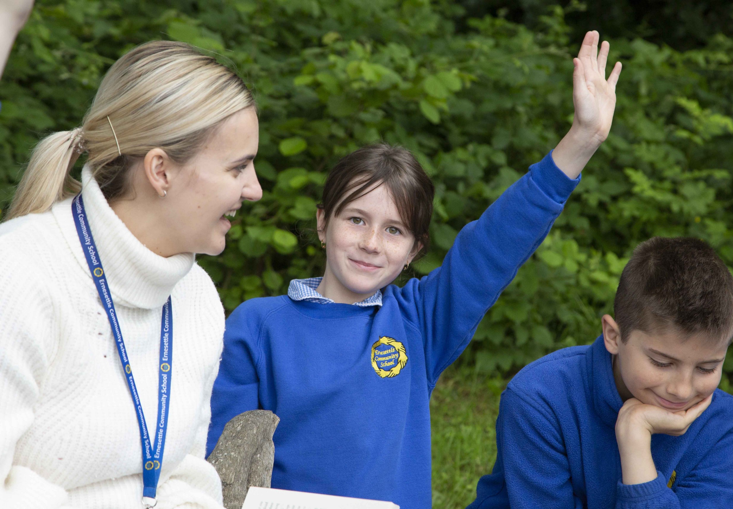 two pupils sat working with a teacher in a green outdoor space